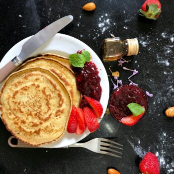 Top view of pancakes with strawberry jam, fresh strawberries, almonds, and honey on a dark kitchen counter.