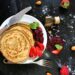 Top view of pancakes with strawberry jam, fresh strawberries, almonds, and honey on a dark kitchen counter.