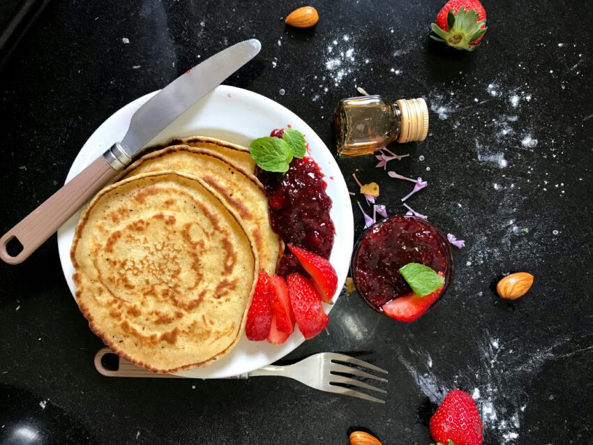 Top view of pancakes with strawberry jam, fresh strawberries, almonds, and honey on a dark kitchen counter.