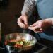 Chef seasoning a vegetable stir fry in a pan with fresh herbs on a stove.