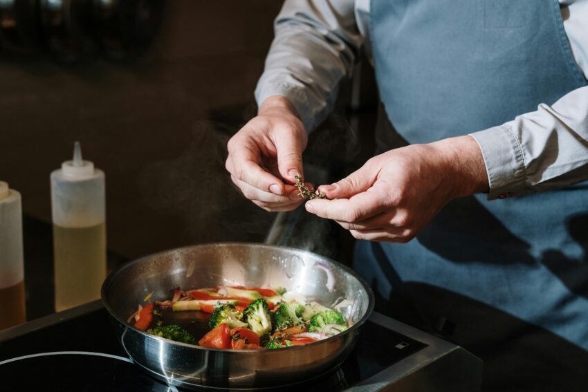 Chef seasoning a vegetable stir fry in a pan with fresh herbs on a stove.